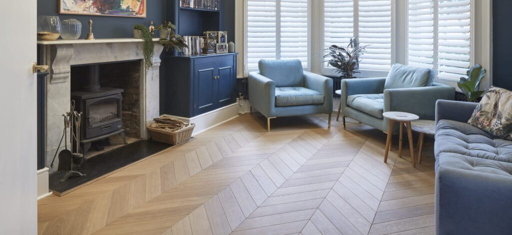 A cozy living room with blue armchairs, a blue sofa, wooden chevron flooring, a fireplace, and shuttered windows. Potted plants and decor are placed around the room.