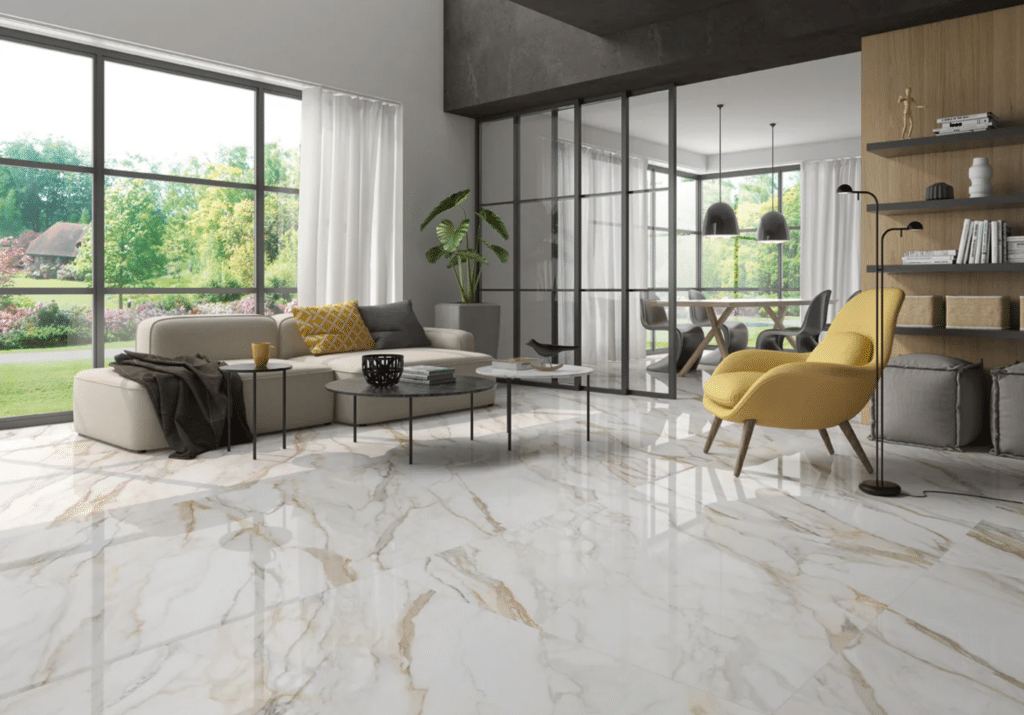 Modern living room with large marble tile floor, light gray sofa, yellow chair, coffee table, and glass wall separating dining area; large windows show a green outdoor garden.